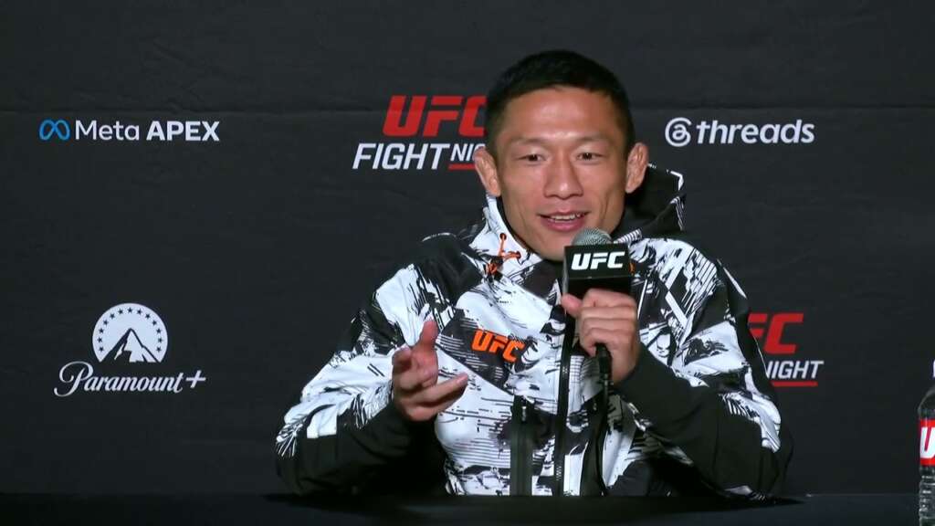 MMA fighter speaks at a UFC press conference, holding a microphone and gesturing, wearing a black-and-white patterned jacket with a UFC logo.