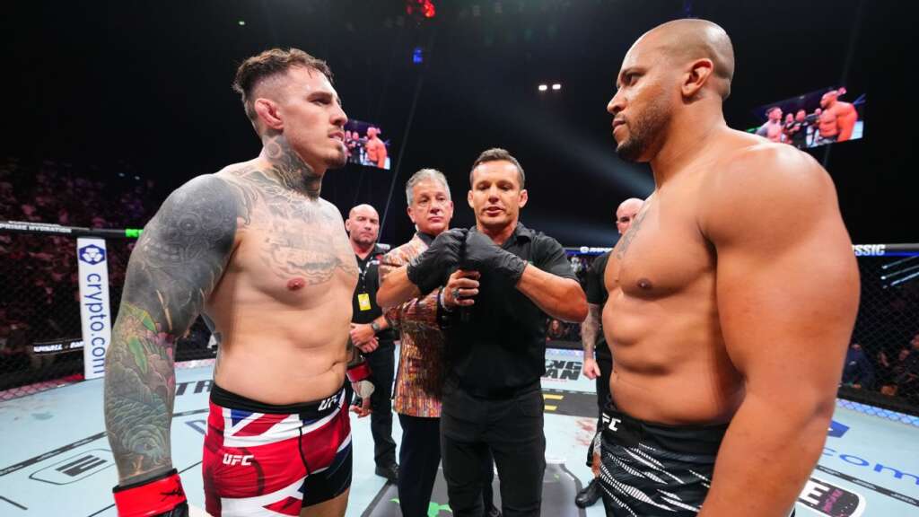 Two shirtless UFC fighters face off in the octagon, with a referee between them and a cheering crowd around the cage.