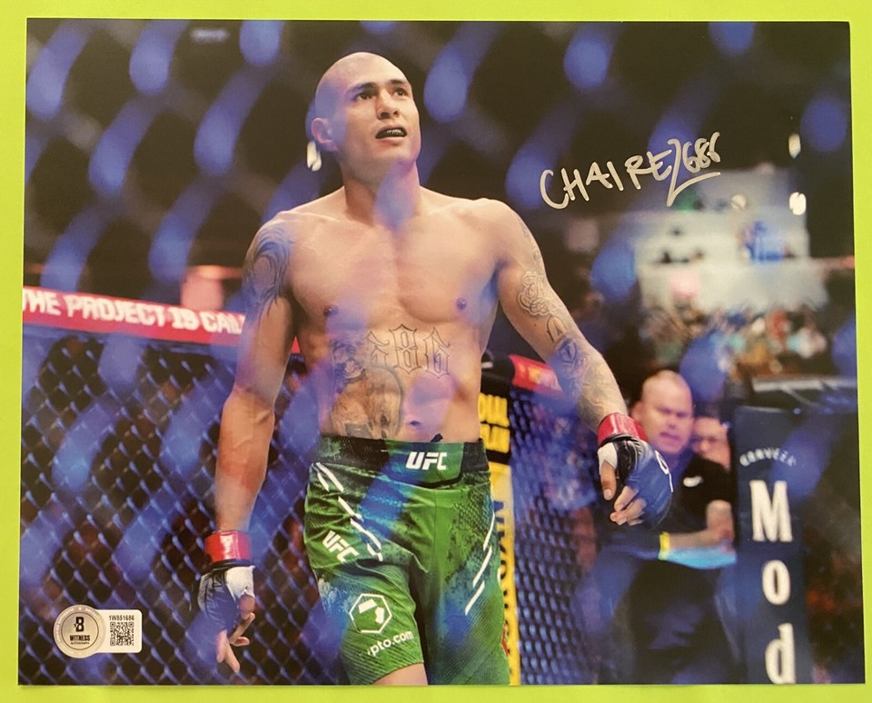 Male mixed martial artist in the UFC octagon, shirtless with green shorts and red gloves, tattoos visible, standing with crowd behind him.