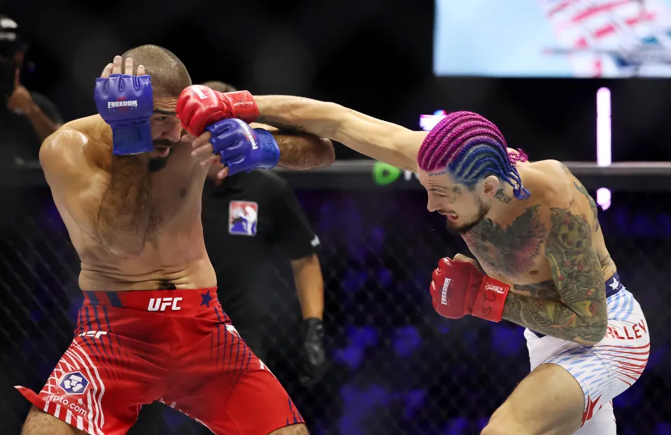 Two UFC fighters exchange punches in the octagon, one in red shorts and blue gloves, the other with colorful braids and tattoos delivering a punch.