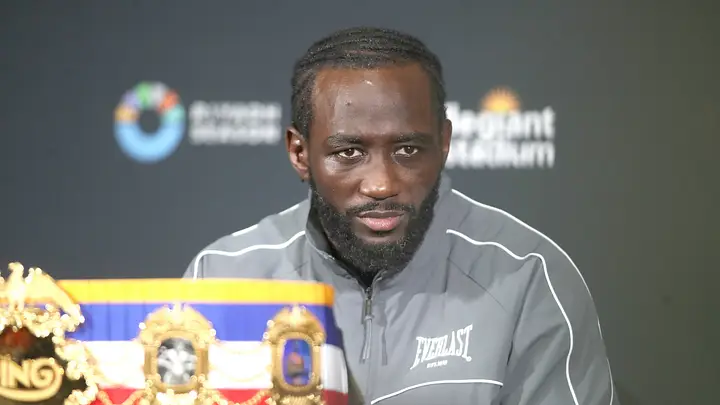 Bearded man in a gray Everlast jacket at a press conference, sitting behind a boxing championship belt on a table.