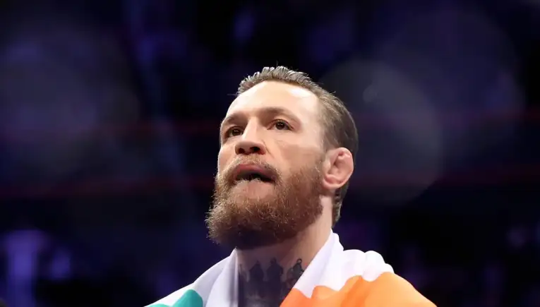 Close-up of a bearded man in a boxing ring, wrapped in an Irish flag, looking up with a focused expression.