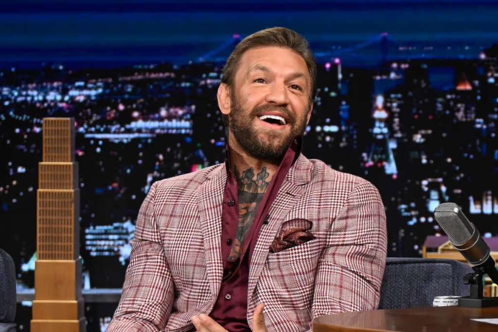 Man in a burgundy check blazer sits on a talk-show set, laughing with a cityscape backdrop behind him.