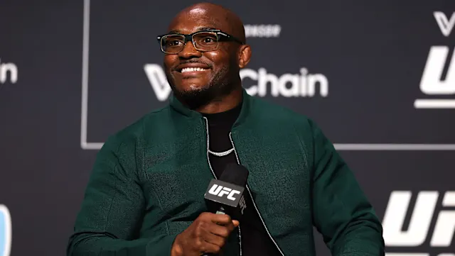 0bb79332599b43de4fcb2b8b0aeb691e - AfroCombat Smiling man in a green jacket speaks into a UFC microphone at a press event, with sponsor logos in the background.