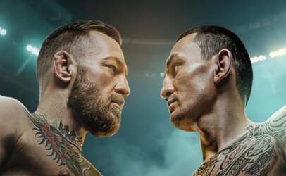 Two tattooed men face off shirtless in a stadium, intense stare-down under bright arena lights.