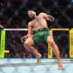 UFC fighter in green shorts kicking mid-air inside the octagon during a match, with yellow ropes and crowd in the background.