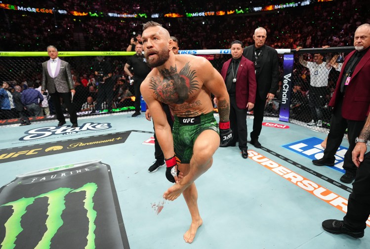 MMA fighter with green shorts and prominent chest tattoos steps out of the cage as officials and a cheering crowd look on.
