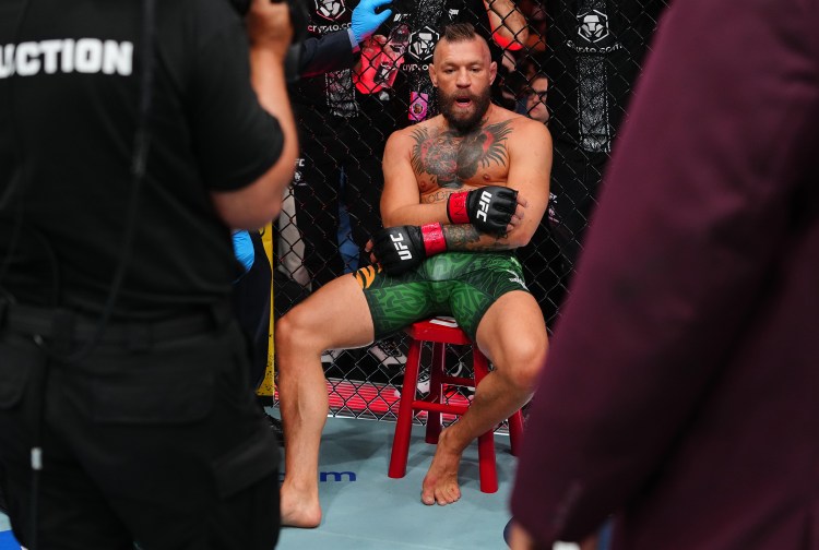 UFC fighter with tattoos sits on a red stool in the cage, arms crossed, wearing green shorts and red gloves.