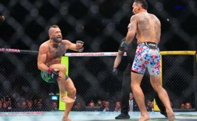 Two shirtless male fighters clash inside a fenced MMA cage, one delivering a kick as the other faces him, crowd watching in the background.