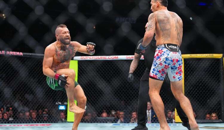 Two shirtless male fighters clash inside a fenced MMA cage, one delivering a kick as the other faces him, crowd watching in the background.