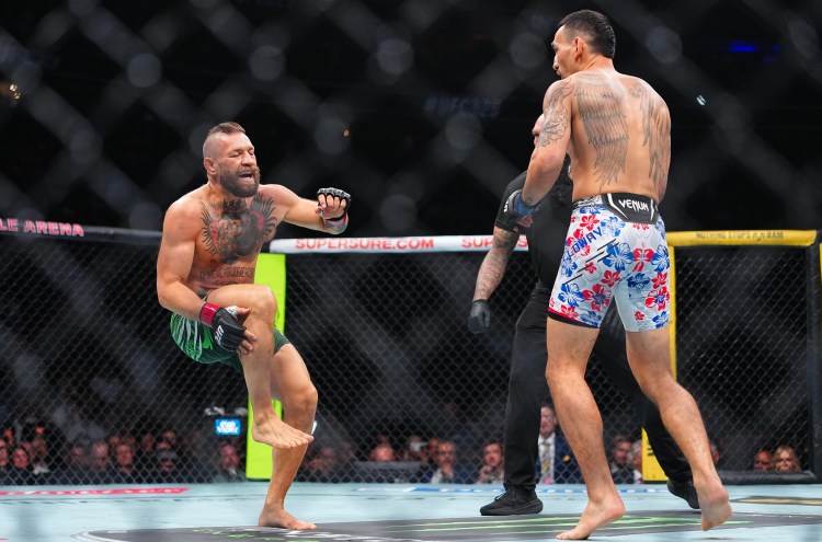 Two shirtless male fighters clash inside a fenced MMA cage, one delivering a kick as the other faces him, crowd watching in the background.