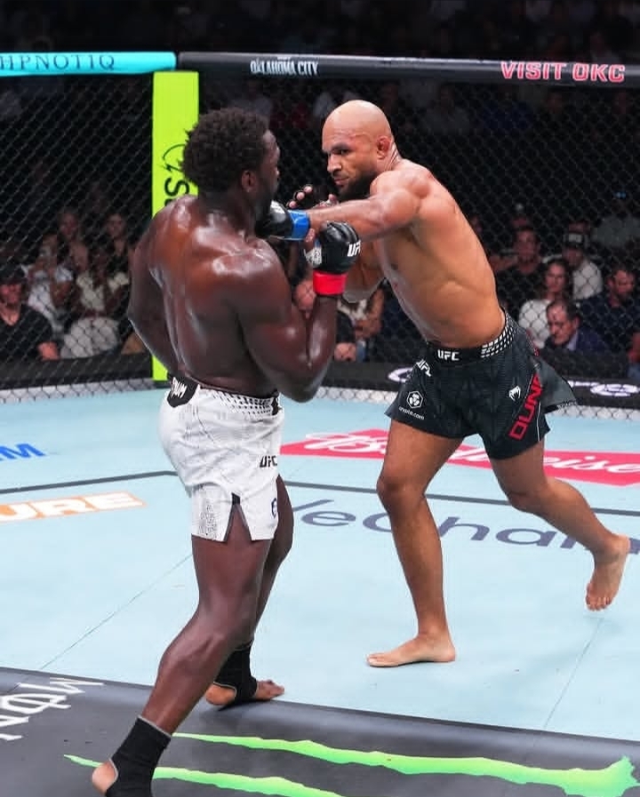 Oplus_131072 - AfroCombat Two UFC fighters clash in the octagon as one lands a punch on the other; the bald fighter wears black shorts and the opponent wears white shorts, with a cheering crowd behind them.