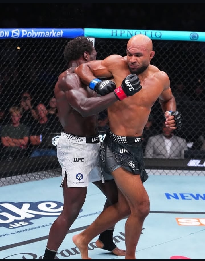 Oplus_131072 - AfroCombat Two UFC fighters in a cage exchange clinch punches during a match; one fighter in white shorts, the other in dark shorts, both wearing UFC gloves as spectators watch from outside the fence.