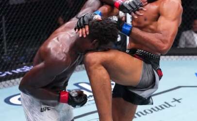 Two male MMA fighters clash in a cage; one delivers a knee to the other's midsection while they grapple in close quarters, wearing MMA gloves and trunks.