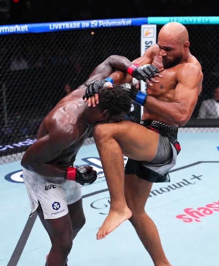 Oplus_131072 - AfroCombat Two male MMA fighters clash in a cage; one delivers a knee to the other's midsection while they grapple in close quarters, wearing MMA gloves and trunks.