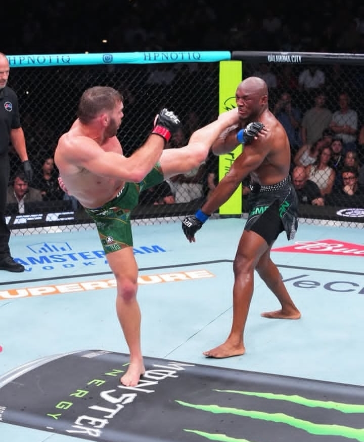 Oplus_131072 - AfroCombat MMA fight in a cage: left fighter in green shorts lands a high kick on the opponent in black shorts with blue gloves; audience watches in the background.