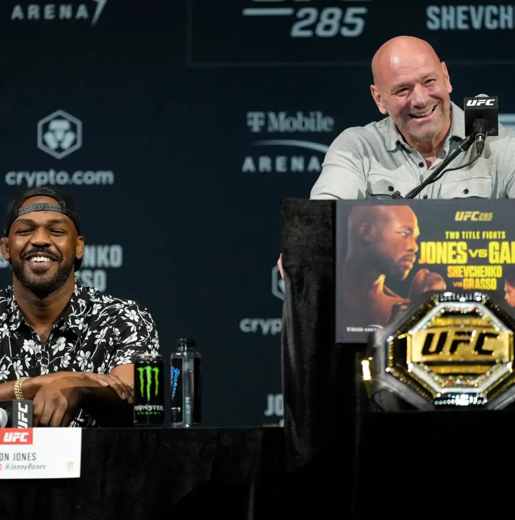 Smiling bald man at a UFC press conference, speaking into a microphone with a promotional poster at the podium and sponsor logos in the background.
