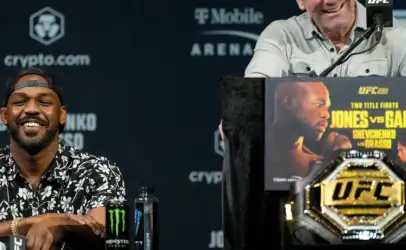 Smiling bald man at a UFC press conference, speaking into a microphone with a promotional poster at the podium and sponsor logos in the background.