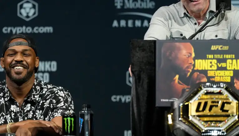 Smiling bald man at a UFC press conference, speaking into a microphone with a promotional poster at the podium and sponsor logos in the background.