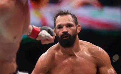 Intense moment of a shirtless, bearded mixed martial artist sweating in the ring, facing an opponent's gloved fist near his face.