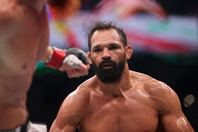 Intense moment of a shirtless, bearded mixed martial artist sweating in the ring, facing an opponent's gloved fist near his face.