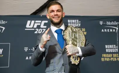 UFC fighter in a gray plaid suit posing with a gold championship belt draped over his shoulder, smiling at the camera while raising one finger in victory against a UFC 207 backdrop.