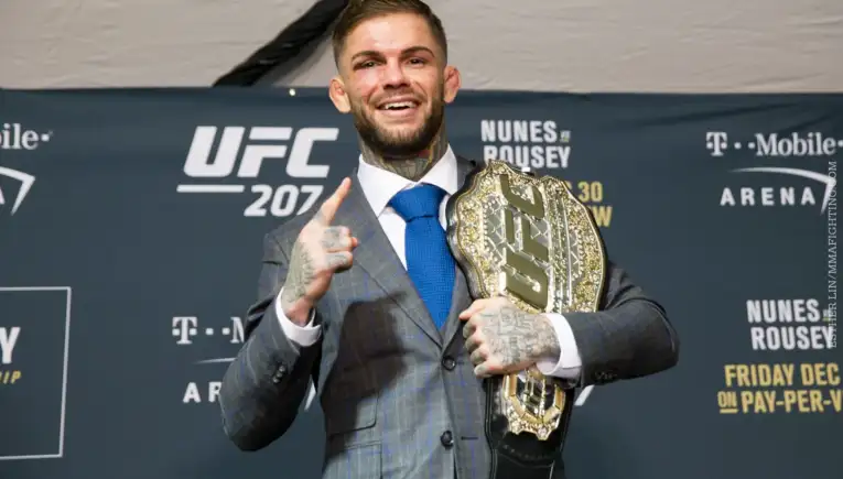 UFC fighter in a gray plaid suit posing with a gold championship belt draped over his shoulder, smiling at the camera while raising one finger in victory against a UFC 207 backdrop.