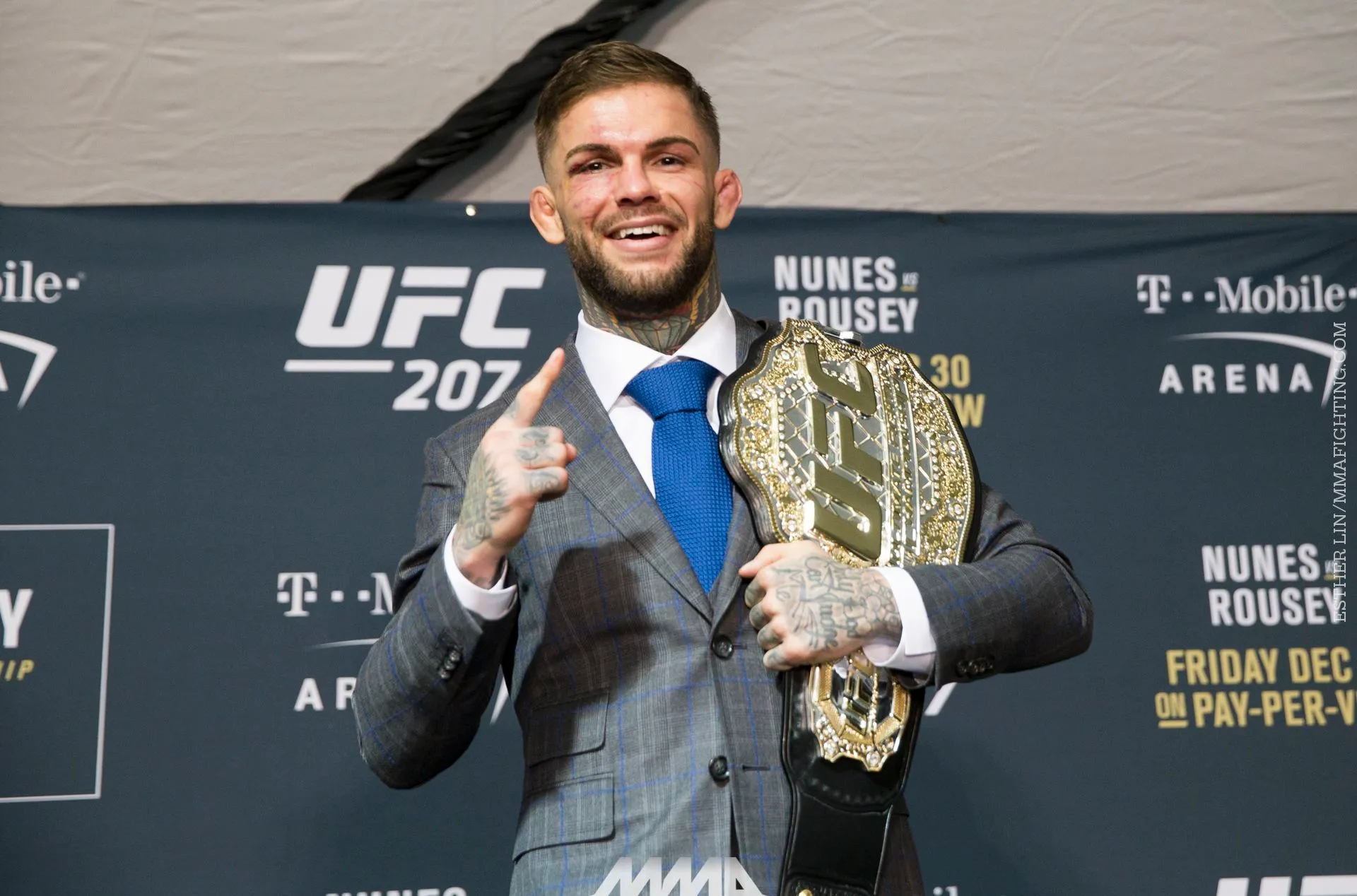 UFC fighter in a gray plaid suit posing with a gold championship belt draped over his shoulder, smiling at the camera while raising one finger in victory against a UFC 207 backdrop.