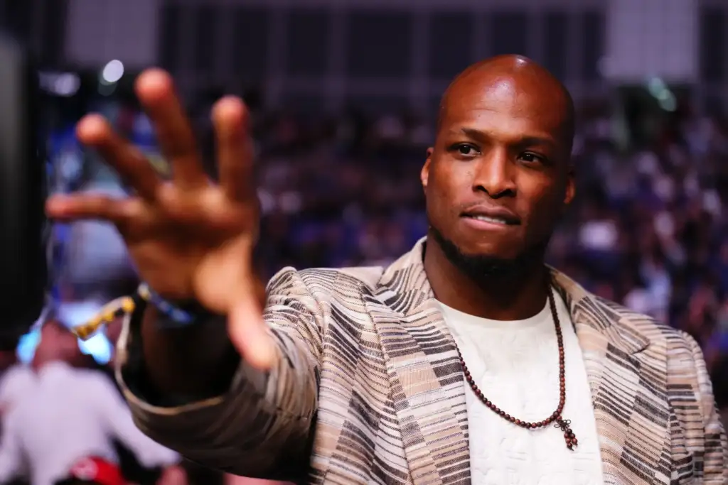 1563540121 - AfroCombat Man with a striped jacket and beaded necklace taking a selfie at a crowded event, reaching toward the camera.