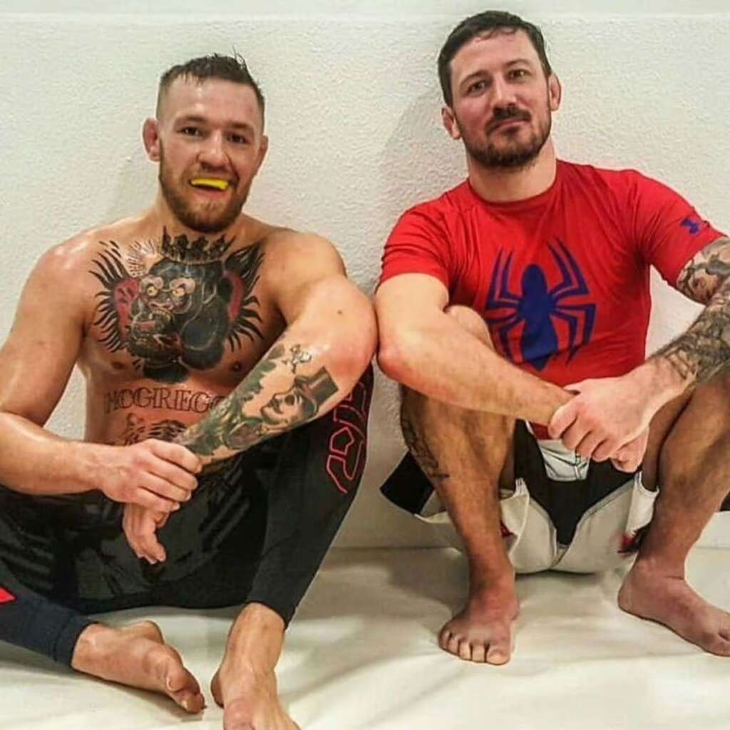 Two tattooed men sit on a gym mat against a white wall, smiling; one is shirtless with a chest and arm full of ink and a yellow mouthguard, the other wears a red Spider-Man shirt.