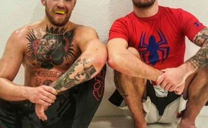 Two tattooed men sit on a gym mat against a white wall, smiling; one is shirtless with a chest and arm full of ink and a yellow mouthguard, the other wears a red Spider-Man shirt.