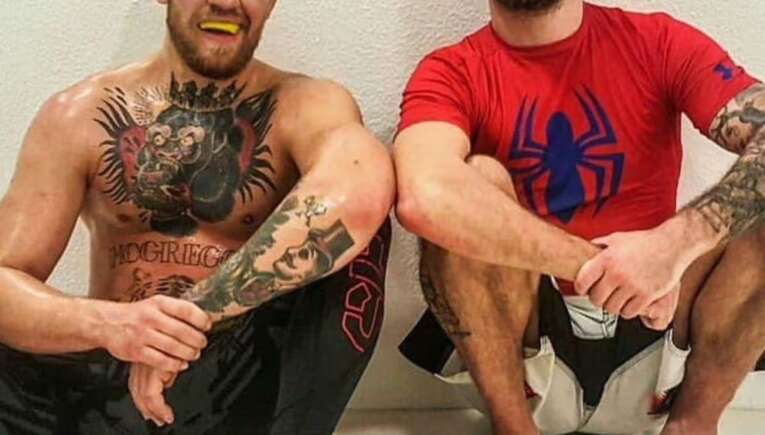 Two tattooed men sit on a gym mat against a white wall, smiling; one is shirtless with a chest and arm full of ink and a yellow mouthguard, the other wears a red Spider-Man shirt.
