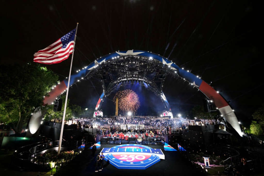 Nighttime concert under a large arch stage with fireworks, a waving American flag, and a large audience.