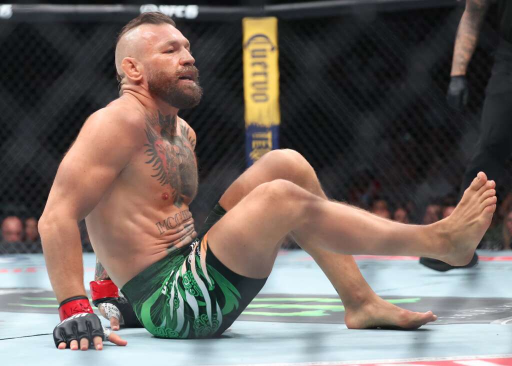 Bearded MMA fighter with a mohawk sits on the mat inside a cage, wearing green patterned shorts and red wrist wraps.