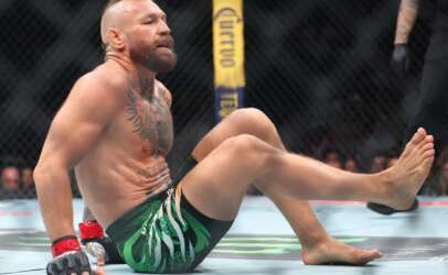 Bearded MMA fighter with a mohawk sits on the mat inside a cage, wearing green patterned shorts and red wrist wraps.