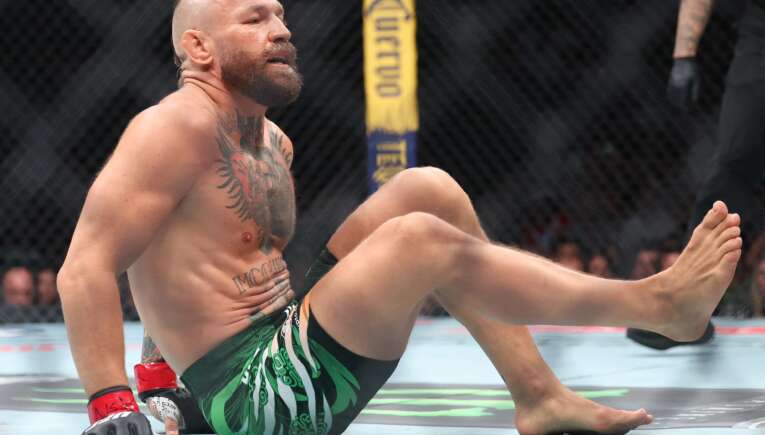 Bearded MMA fighter with a mohawk sits on the mat inside a cage, wearing green patterned shorts and red wrist wraps.