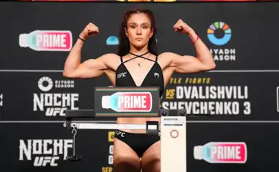 Female UFC fighter on a weigh-in stage, arms raised in a double biceps pose, wearing a black athletic outfit with a scale in front and event banners in the background.