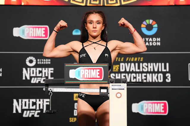 Female UFC fighter on a weigh-in stage, arms raised in a double biceps pose, wearing a black athletic outfit with a scale in front and event banners in the background.