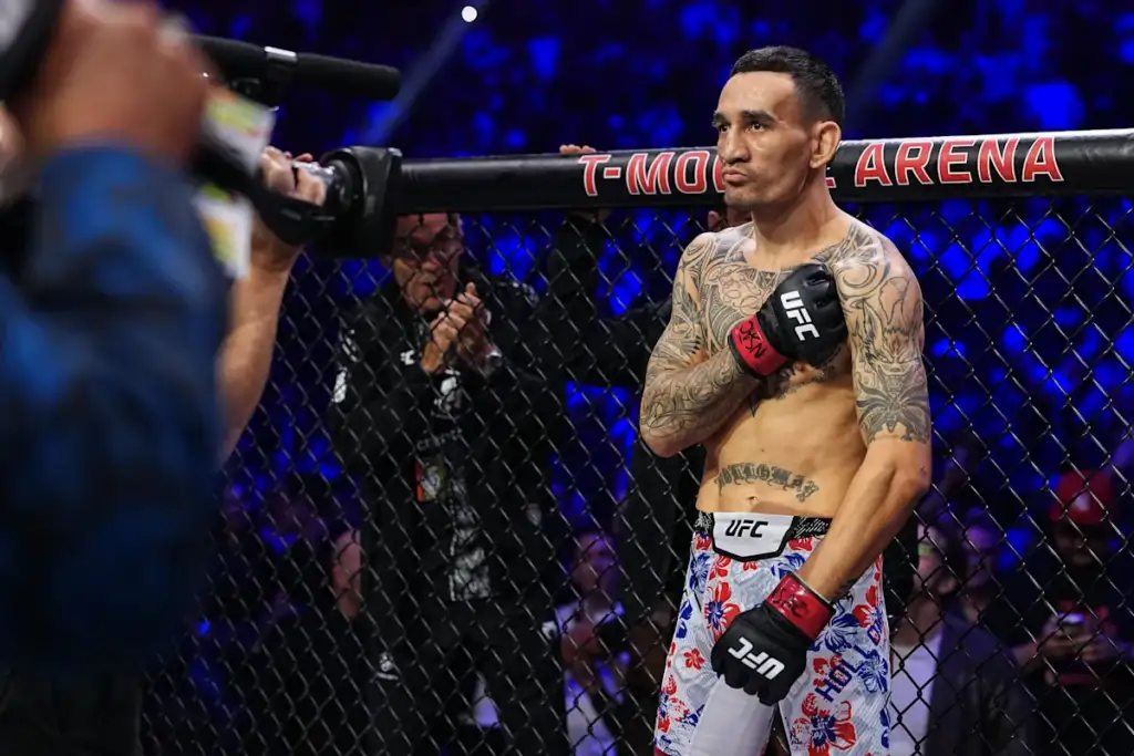 UFC fighter standing inside a cage, wearing floral shorts and red gloves, leaning on the fence as a camera records nearby.