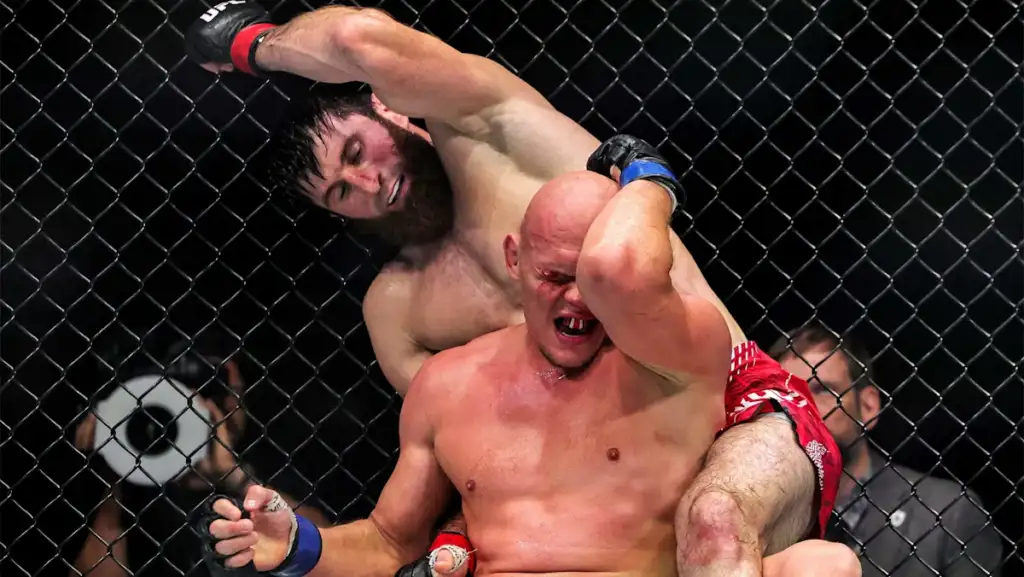 MMA fight inside a chain-link cage; a wrestler in red shorts is held in a rear headlock by a opponent with black gloves, both in visible effort and pain.