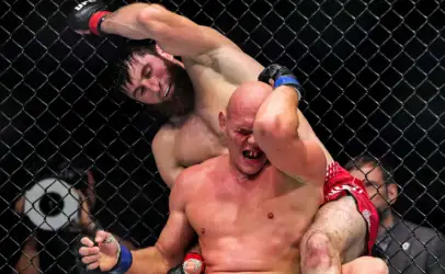 MMA fight inside a chain-link cage; a wrestler in red shorts is held in a rear headlock by a opponent with black gloves, both in visible effort and pain.