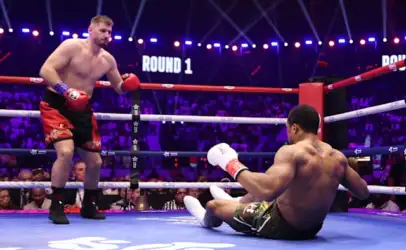 Boxer in red trunks and blue gloves stands over a fallen opponent in white gloves and green shorts on the canvas during Round 1 in a crowded boxing arena.