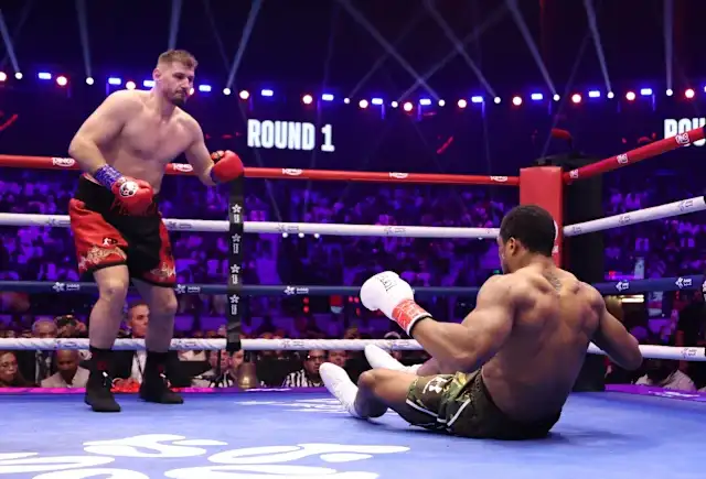 Boxer in red trunks and blue gloves stands over a fallen opponent in white gloves and green shorts on the canvas during Round 1 in a crowded boxing arena.
