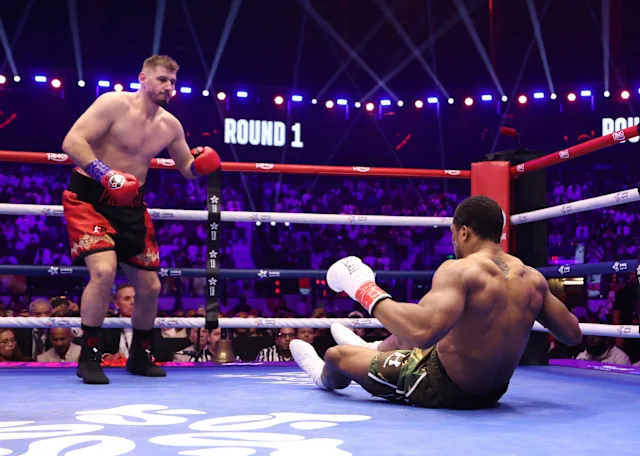 Boxer in red trunks and blue gloves stands over a fallen opponent in white gloves and green shorts on the canvas during Round 1 in a crowded boxing arena.
