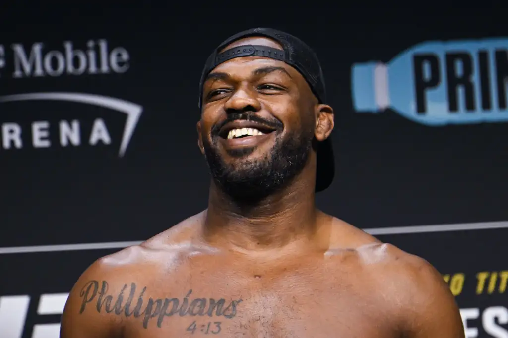 Smiling shirtless man wearing a backward black cap, with a tattooed chest reading 'Philippians 4:13' seen in front of a stage backdrop.