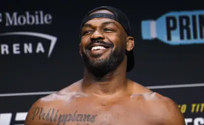 Smiling shirtless man wearing a backward black cap, with a tattooed chest reading 'Philippians 4:13' seen in front of a stage backdrop.