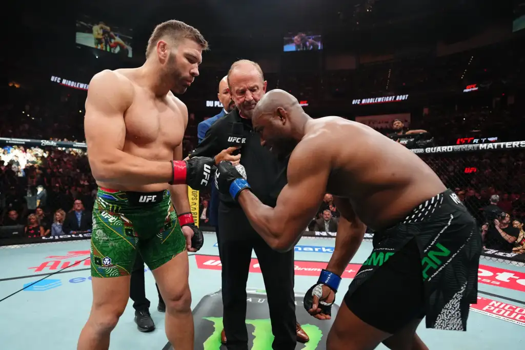 Two UFC fighters stand in the octagon while a referee speaks into a microphone between them, amid a cheering crowd.
