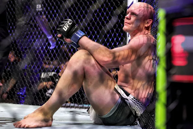 UFC fighter sits exhausted inside the cage corner, knees bent, gloves on, gazing upward as shadows and a crowd loom behind the chain-link fence.
