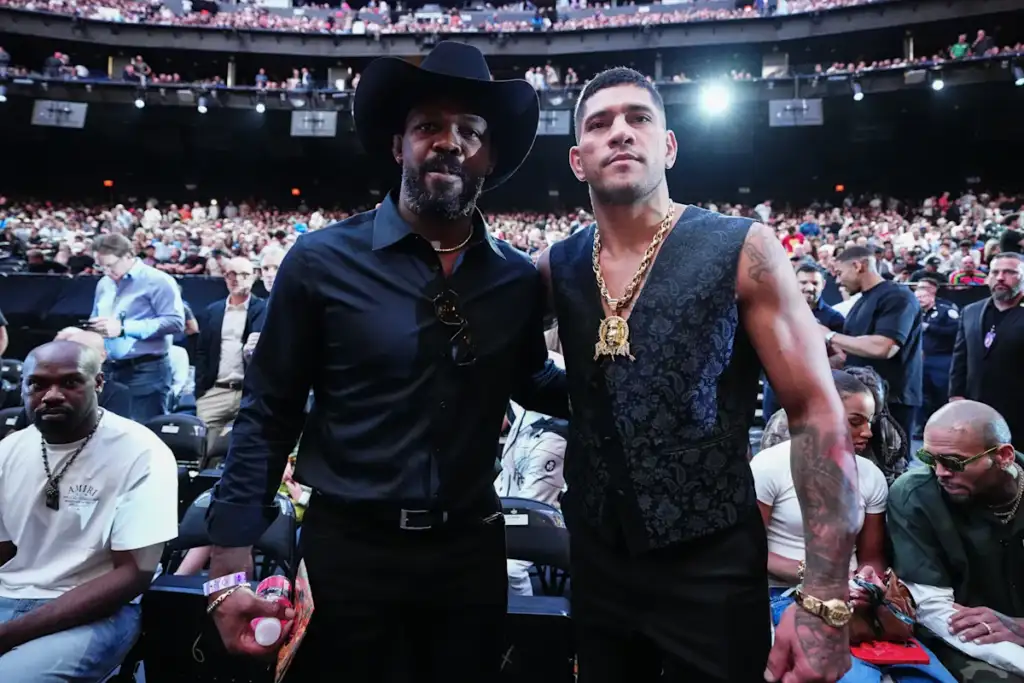 Two men pose for a photo in a crowded arena, one wearing a black cowboy hat and shirt, the other in a dark patterned vest with a gold chain.