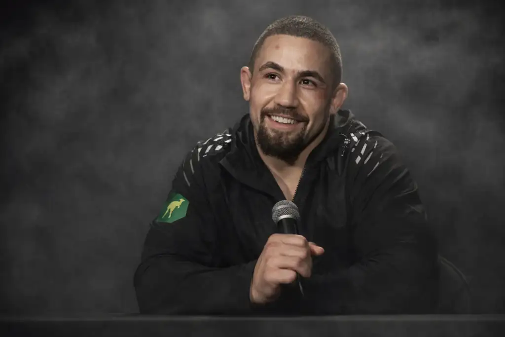 Smiling man in a black jacket holds a microphone at a desk with a smoky gray backdrop behind him.